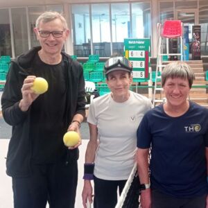 Zwei Frauen stehen links und rechts von einem Tennisnetz. Neben der linken Frau steht noch ein Mann, der Blindentennisbälle in beiden Händen hält. Alle drei Personen lächeln. Sie sind sportlich gekleidet.