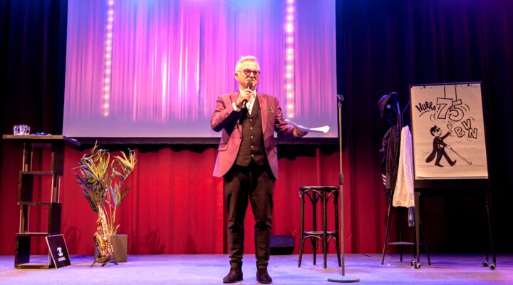 Jörg Knör steht mit einladender Geste auf der Bühne, in seiner linken Hand das Mikro. Er hat helle graue Haare, trägt eine Brille, über einem weißen Hemd und dunkler Weste einen helleren Blazer, dazu eine dunkle Hose