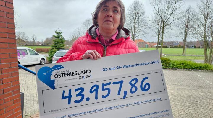 Eine ältere Frau hält einen großen Scheck in die Höhe. Auf diesem steht die Summe von 43957,86 €. Die Frau ist blind. Im Hintergrund ist ein Parkplatz mit Bäumen zu sehen.