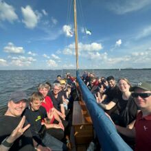 Circa 20 Personen sitzen gemeinsam in einem langen Boot. Sie zeigen mit ihrer Hand eine Gebärde und lächeln. Im Hintergrund sind Wasser und der blaue Himmel zu sehen.