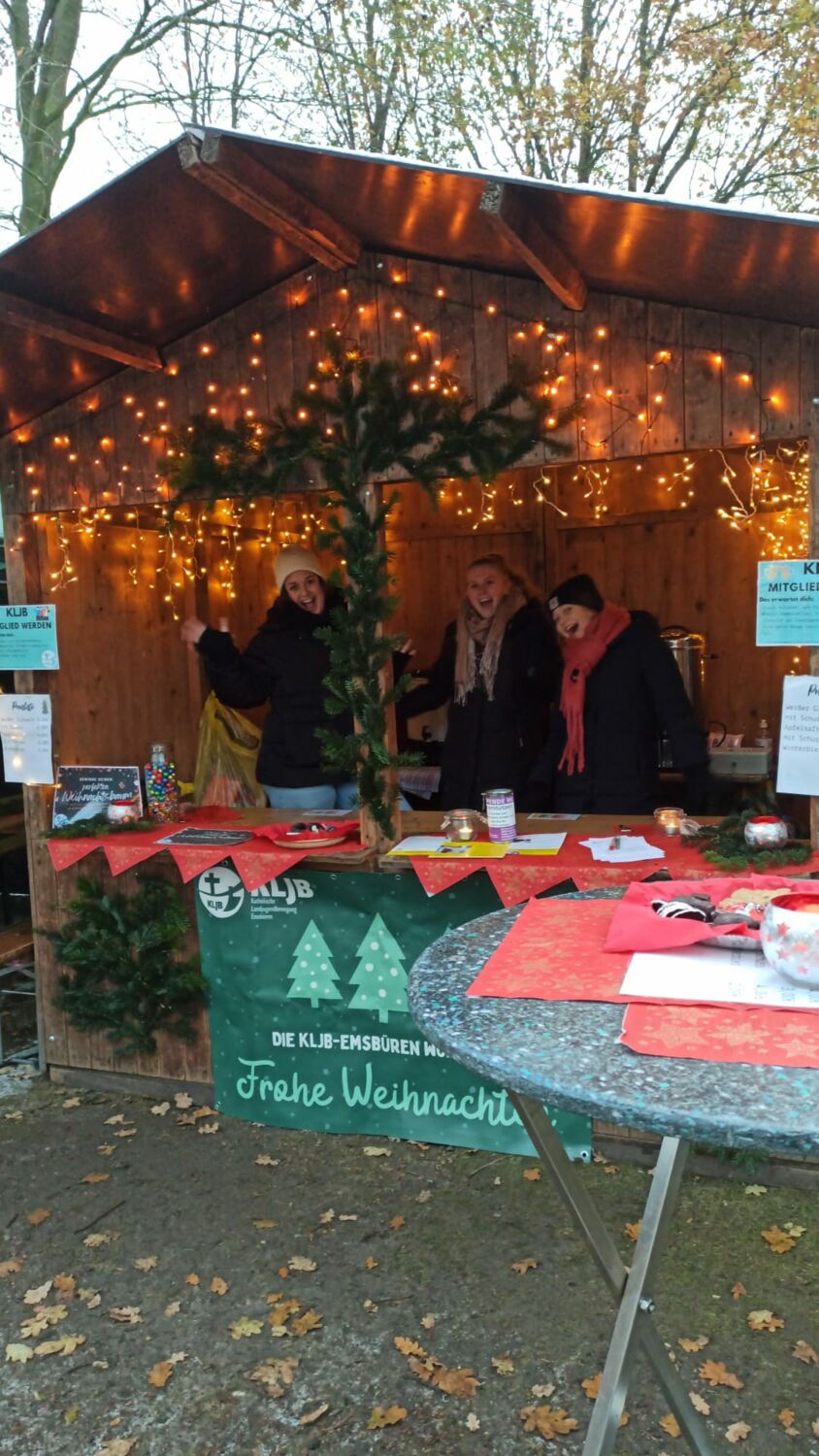 Hinter einem Holzstand auf dem Weihnachtsmarkt stehen drei junge Freuen, die fröhlich lachen und winken. Sie tragen Wintersachen. Auf dem Stand liegen viele Informationsmaterialien.