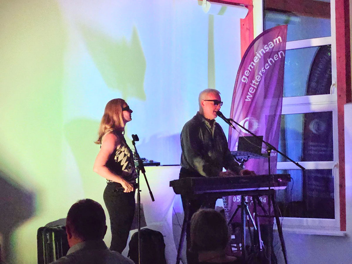 Eine junge Frau und ein älterer Mann mit weißem Haar, beide mit Sonnenbrille, musizieren zusammen. Sie hält ein Mikrofon in der Hand und er spielt singend am Piano.